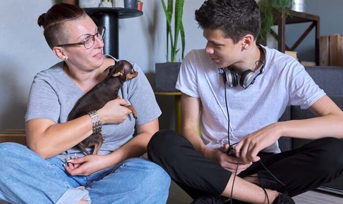 Happy talking mom and teenage son, parent and child sitting at home on the floor Happy talking mom and teenage son, parent and child sitting at home on the floor with pet little dog toy terrier, real friendly loving family